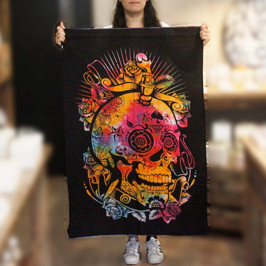 A person holds a tapestry depicting a colorful, ornate skull design against a dark background. The skull is surrounded by flowers and other decorative elements. The context is an indoor setting.