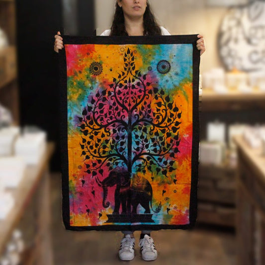 A person holds a tie-dye tapestry depicting a black elephant beneath a stylized tree. The tapestry shows vibrant rainbow colors and is displayed in a shop.