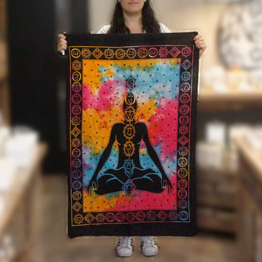 A person holds a tie-dye tapestry depicting a meditating figure with chakra symbols; a blurred shop background is visible.