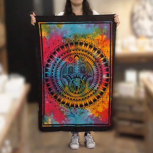 A person holds a tie-dye tapestry featuring a hamsa hand inside a mandala. The vibrant colors are predominantly pink, orange, yellow, and blue. The background is a blurred shop interior.