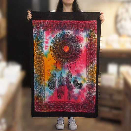 A person displays a tie-dye tapestry featuring a dreamcatcher design. The tapestry is predominantly pink, orange, and blue with black detailing. The background is a blurred shop interior.