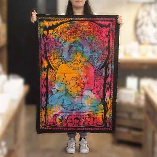A tie-dye tapestry depicting a meditating Buddha is held by a person. The tapestry features vibrant colors and intricate detailing. The context is an indoor shop setting.