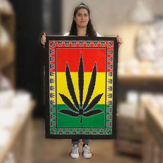 A woman holds a tapestry depicting a marijuana leaf on a red, yellow, and green background, in a blurred retail setting.
