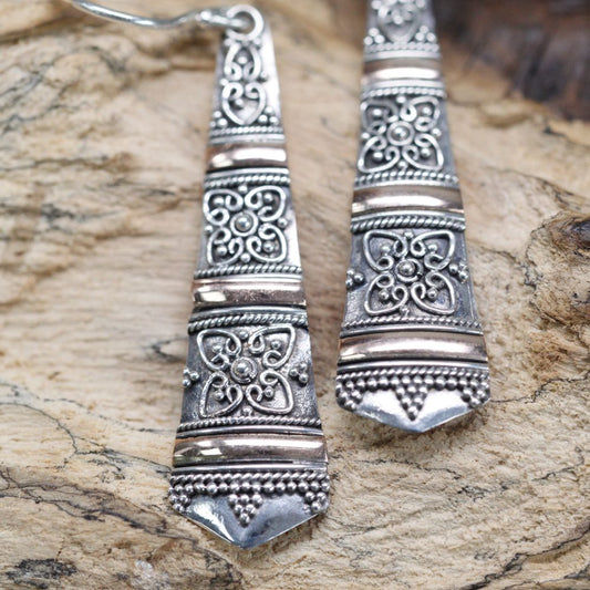 Two silver earrings with ornate designs rest on a textured, light brown wood surface. The earrings are elongated and taper slightly towards the bottom.