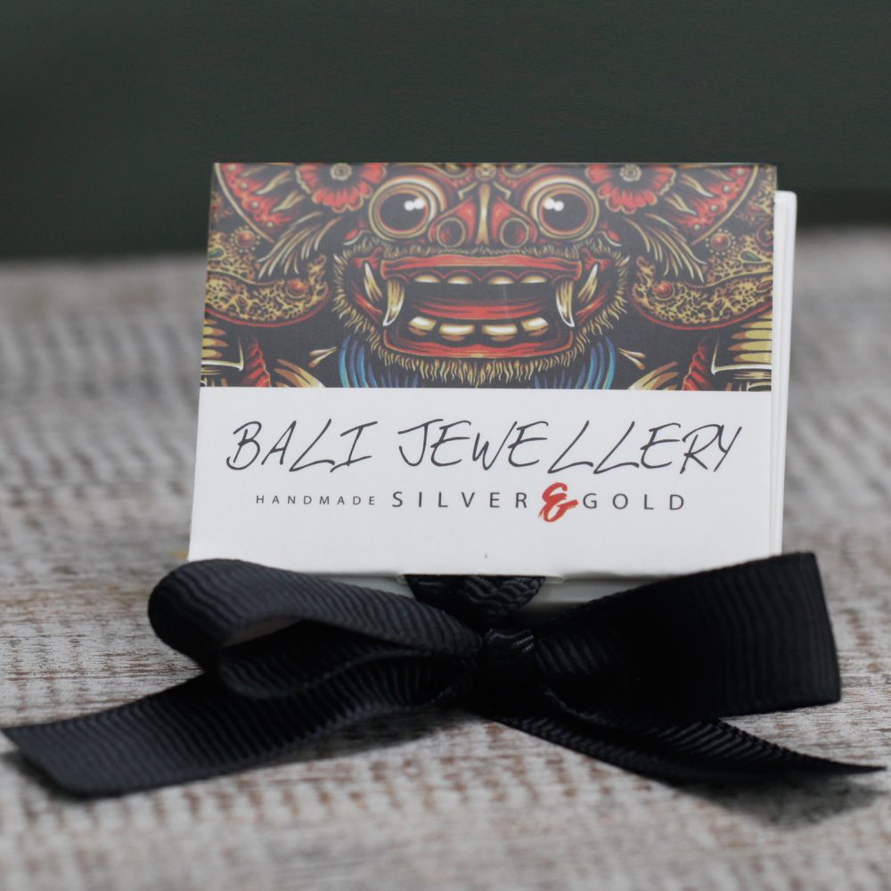 A "BALI JEWELLERY HANDMADE SILVER & GOLD" tag is tied with a black ribbon. It sits on a light gray surface, and above the tag is a colorful illustration.