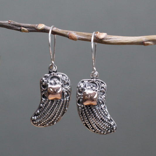 Two silver earrings hang from a twig. The earrings have curved, textured bodies with small, gold squares near the top. The background is a soft gray.