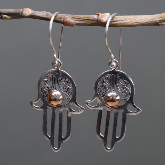 Two silver hamsa earrings hang from a twig. Each earring has a copper-colored bead in the palm. The background is a gray color.