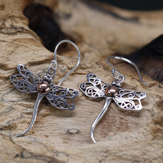 Two silver dragonfly earrings rest on a textured, light-brown piece of wood. The earrings have copper-colored bodies and ornate, cutout wings.