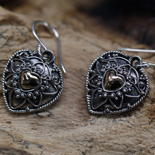 Two ornate, heart-shaped earrings, detailed with silver filigree and a central gold heart, rest on a rough-textured, light brown wooden surface. The earrings dangle from silver hooks.