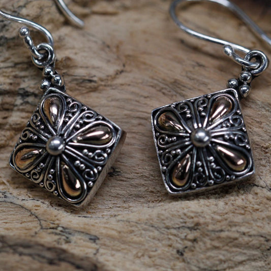 Two silver earrings with floral, teardrop patterns and a small round center rest on a piece of distressed wood. The earrings dangle from curved hooks.