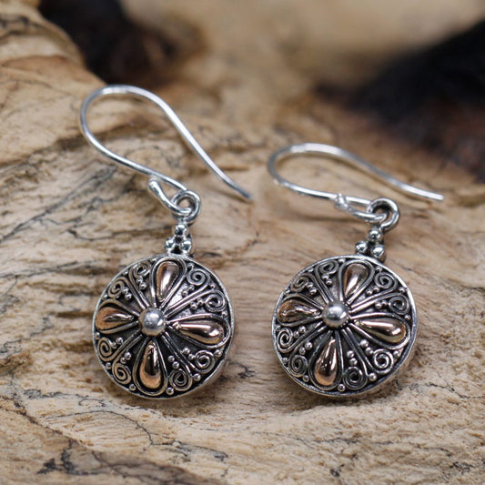 Two ornate, circular silver earrings with a gold floral design dangle on a textured, light brown wooden surface.
