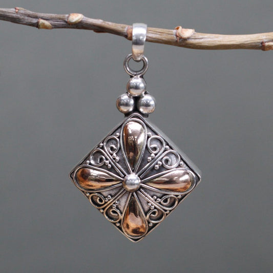 A silver pendant hangs from a twig. The pendant is diamond shaped with gold flower design, a loop for a chain and three stacked silver beads attached at the top.