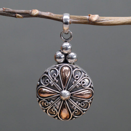 An ornate silver and copper pendant hangs from a small tree branch. The pendant has a decorative flower design and several silver balls at the top.