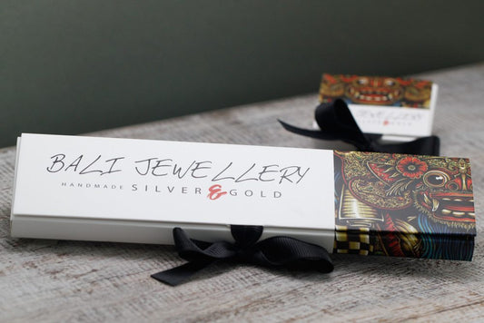 A jewelry box with the text "BALI JEWELLERY, HANDMADE SILVER & GOLD" sits tied with a black bow on a wooden surface. Another, partially visible box with the text "JEWELLERY" is in the background.