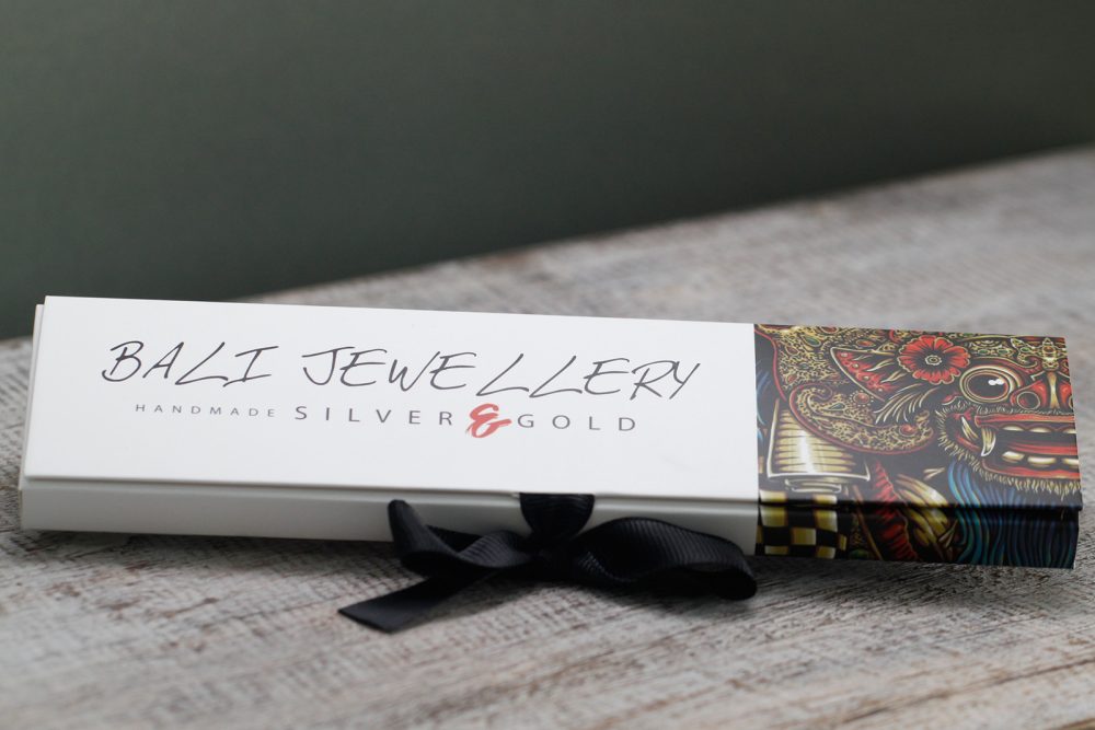 A white box labeled "BALI JEWELLERY HANDMADE SILVER & GOLD" with an ornate, colorful design on one side rests on a gray surface. A black ribbon is tied around the box.