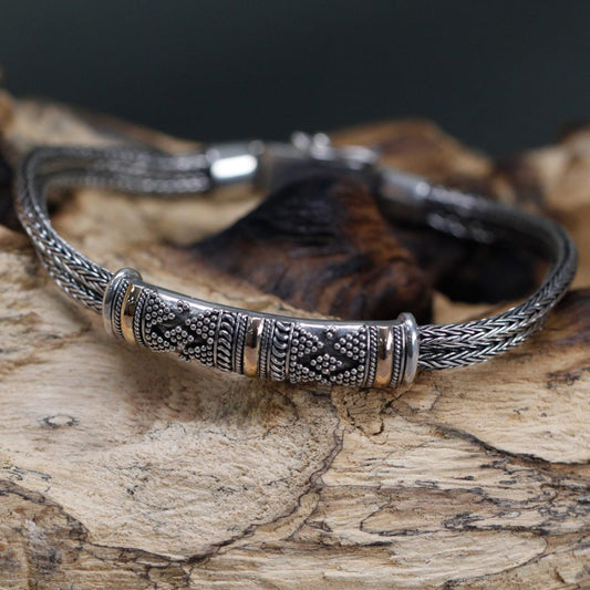 A silver bracelet featuring decorative gold accents and braided strands rests on a piece of driftwood. The bracelet’s ornate central band is decorated with repeating patterns of dots and lines.