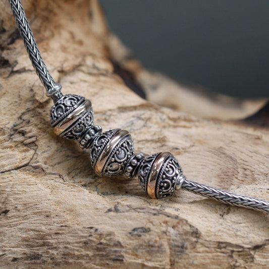 A silver chain with three decorative, filigreed beads rests diagonally on a textured, light brown, wooden surface. The background is blurry, and colored grey.