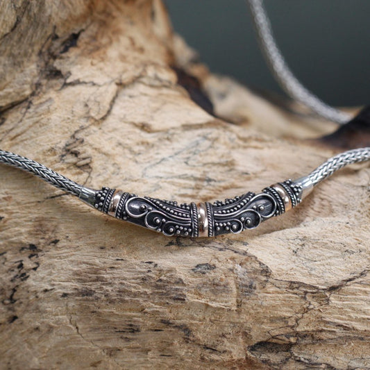 A braided silver necklace with gold-accented beads rests on a piece of textured driftwood. The detailed ornamentation on the central silver piece includes swirls and dots.