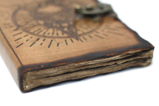 A closed, tan leather journal lies on a white surface. An etched compass design adorns the cover, which is kept shut with a small metal clasp.