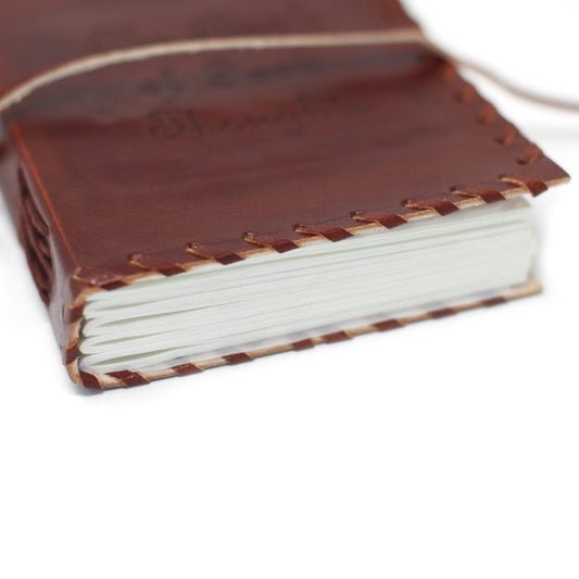A brown leather-bound journal, held closed with twine, sits on a white surface. Visible white pages fill the journal.