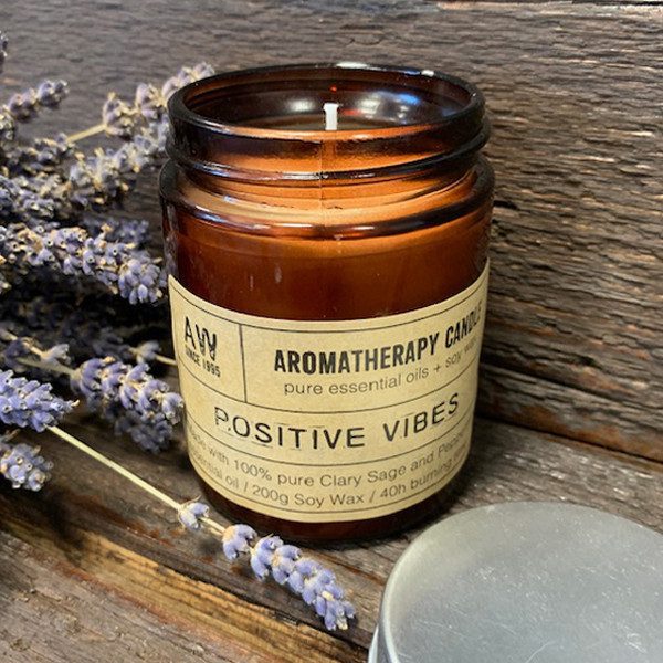 The aromatherapy candle sits on a wooden surface, next to sprigs of lavender. The candle's label reads: "AROMATHERAPY CANDLE pure essential oils + soy wax POSITIVE VIBES Made with 100% pure Clary Sage and Pepper essential oil / 200g Soy Wax / 40h burning time AW SINCE 1995.”