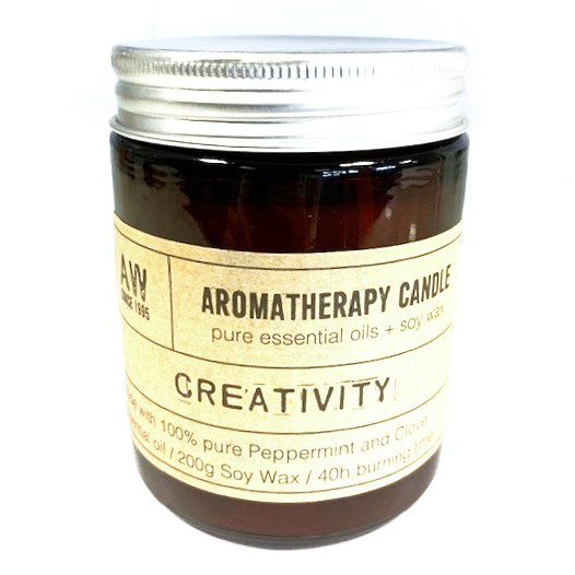 A brown jar candle with a silver lid features a beige label. The label reads: "AW, SINCE 1995, AROMATHERAPY CANDLE pure essential oils + soy wax, CREATIVITY, Made with 100% pure Peppermint and Clove essential oil / 200g Soy Wax / 40h burning time."
