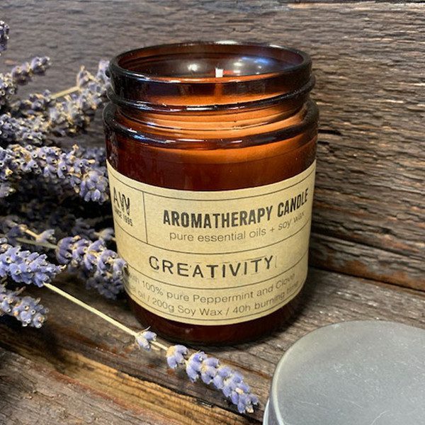 A brown aromatherapy candle labeled "AROMATHERAPY CANDLE Pure essential oils + soy wax CREATIVITY with 100% pure Peppermint and Clove oil / 200g Soy Wax / 40h burning time" sits on a rustic wood surface next to sprigs of lavender.