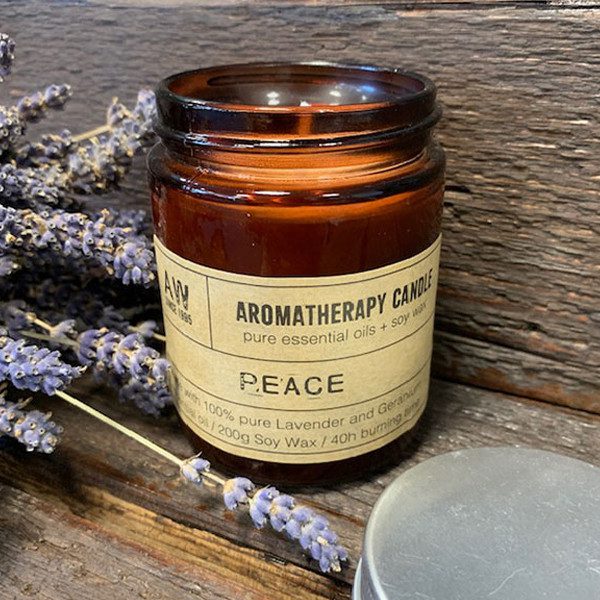 A brown glass candle jar sits on a rustic wooden surface, next to some sprigs of lavender. The jar's label reads: "AROMATHERAPY CANDLE pure essential oils + soy wax PEACE with 100% pure Lavender and Geranium essential oil / 200g Soy Wax / 40h burning time".
