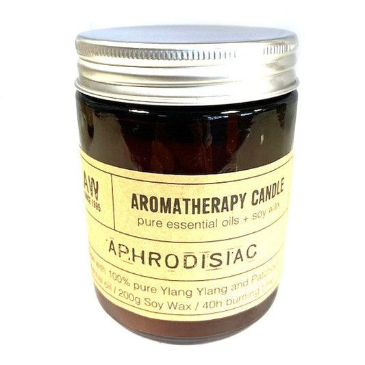 A brown glass aromatherapy candle jar with a silver lid sits on a white surface, labelled “AROMATHERAPY CANDLE pure essential oils + soy wax 'APHRODISIAC' Made with 100% pure Ylang Ylang and Patchouli essential oil / 200g Soy Wax / 40h burning time.”