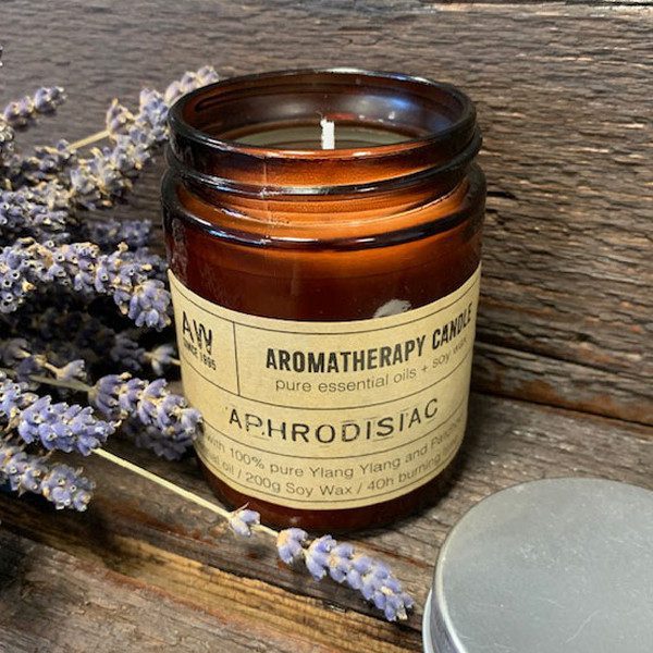 A candle labeled "AROMATHERAPY CANDLE pure essential oils + soy wax APHRODISIAC with 100% pure Ylang Ylang and Patchouli oil / 200g Soy Wax / 40h burning" rests on weathered wood next to sprigs of lavender.