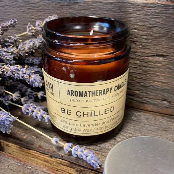 A lit aromatherapy candle sits on a rustic wooden surface next to sprigs of lavender. The label reads: "AWY EST. 1995 AROMATHERAPY CANDLE pure essential oils + soy wax BE CHILLED with 100% pure Lavender and Fennel oil / 200g Soy Wax / 40h burning time".