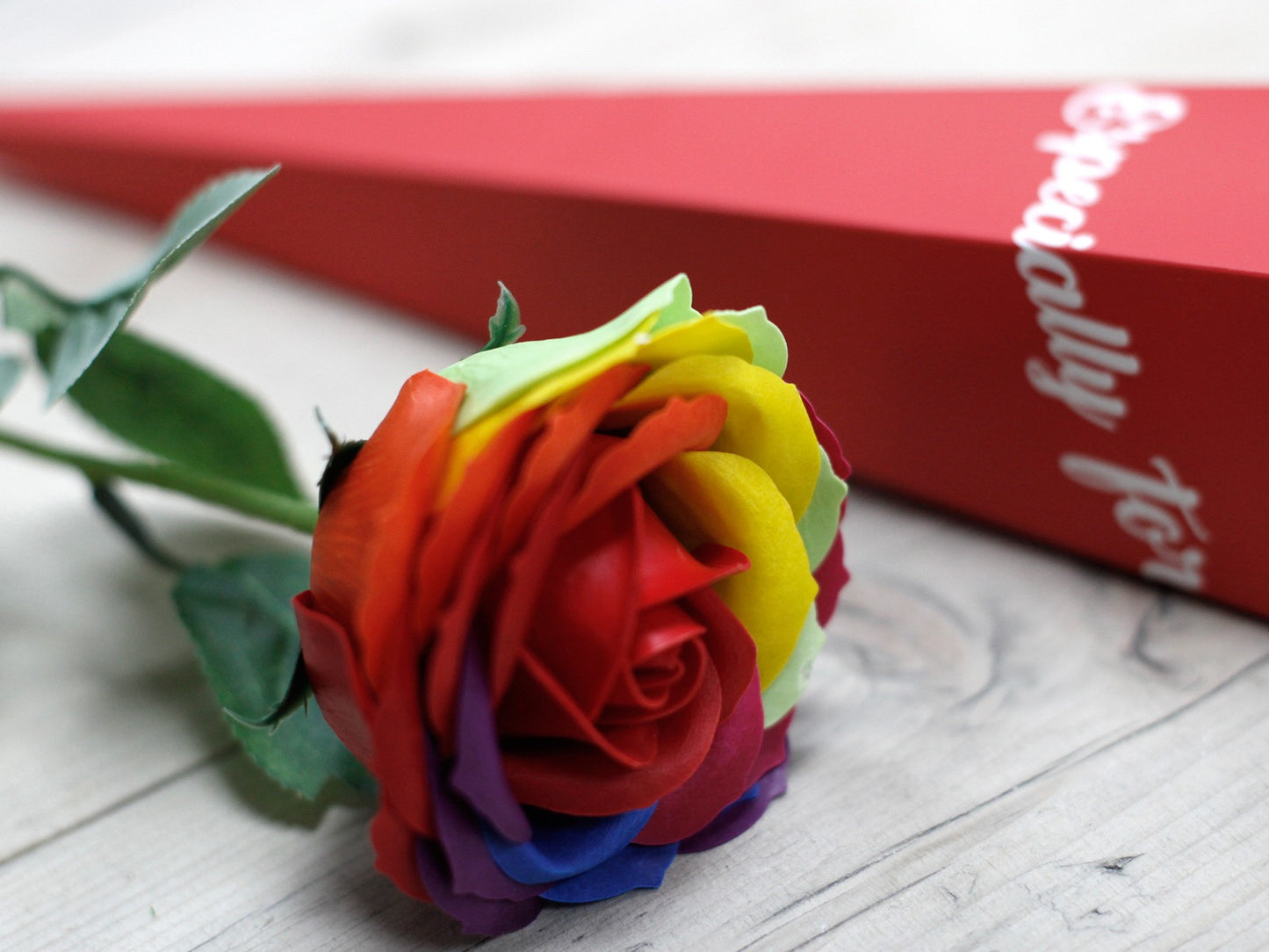 A rainbow rose lies on a wooden surface, next to a red box labeled, "Especially For."