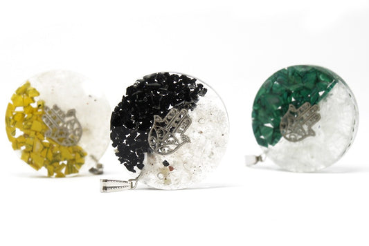Three resin pendants rest on a white surface. The round pendants feature a hamsa hand symbol and are filled with black, green and yellow rock chips.