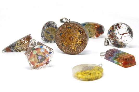 Various orgonite pendants sit on a bright white surface. The pendants have different shapes, with embedded materials, and geometric or chakra symbols.