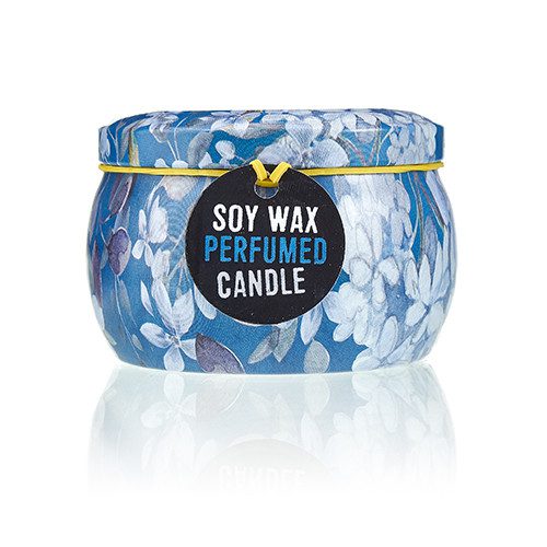 Decorative blue floral tin candle sits on a reflective white surface with a round black tag reading SOY WAX PERFUMED CANDLE and a yellow elastic band securing the lid.