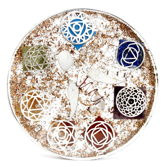 A resin orgone disk is filled with metallic flakes and holds seven chakra symbols, colorful gemstones, two silver wings, and copper coils, placed on a white background.