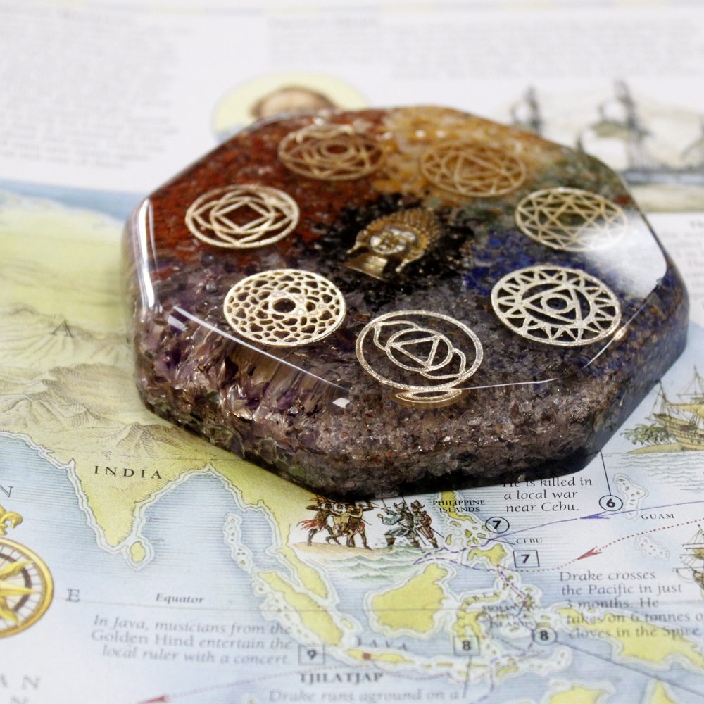 A clear resin orgonite, containing crystals, metal shavings, and chakra symbols, rests on a partially visible map labeled "INDIA" and details about Drake's voyages, including "CEBU" and "TJILATJAP."