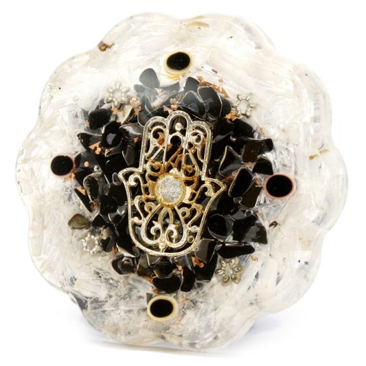 The orgonite amulet features a Hamsa hand at its center, surrounded by black stones, within a translucent, flower-shaped resin mold. It sits against a plain white background.
