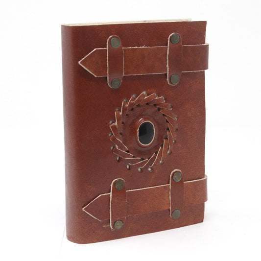 A brown leather journal sits upright. It is decorated with two leather straps and a central circular design with a dark center. The background is plain white.