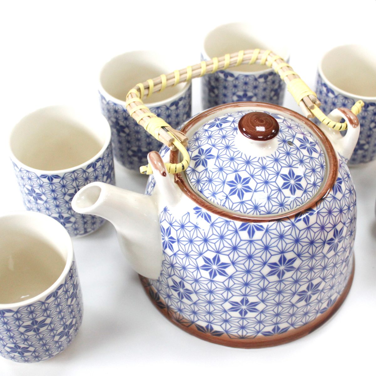 A ceramic teapot with a bamboo handle, adorned with a blue geometric pattern, sits surrounded by six matching teacups on a white surface.