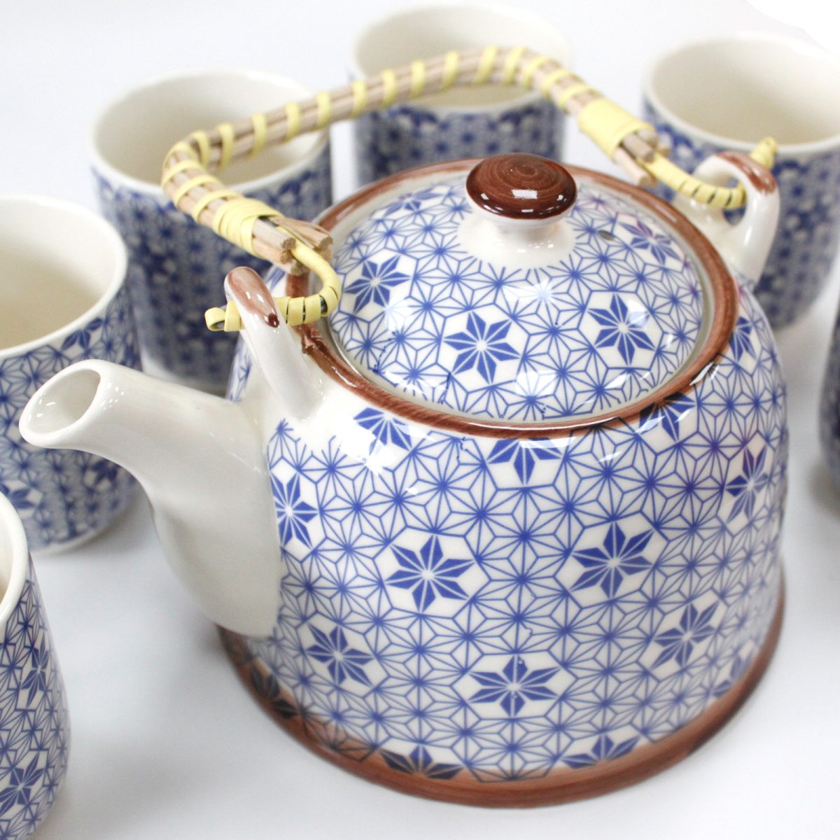 A white ceramic teapot with a blue geometric pattern and a woven handle sits surrounded by matching cups, all arranged on a white surface.