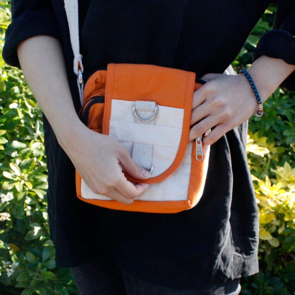 Person holds an orange and white shoulder bag, with a canvas strap, in front of them outdoors, near shrubbery.