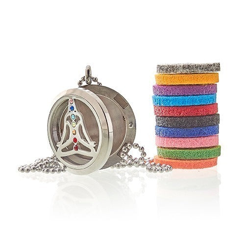 A silver locket with chakra symbols hangs open next to a stack of colorful felt pads. The necklace and pads rest on a bright, reflective surface.