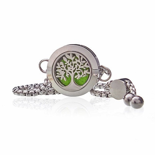A silver locket-style bracelet with a tree-of-life design rests against a white background, casting a reflection below it. A green felt pad is visible behind the tree design.