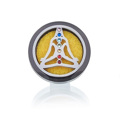 A silver outline of a seated person, marked with seven chakra-colored gems, sits over yellow foam in a round, dark gray locket on a white surface.