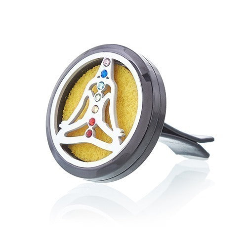 A circular, black car vent diffuser has a silver outline of a meditating person with chakra-colored gemstones in the middle. It rests on a white surface.