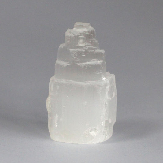 A selenite crystal tower stands against a gray background, its translucent, white form stacked in rough layers that taper to a point.