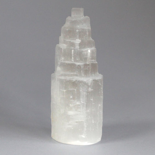 A clear selenite tower stands upright against a neutral gray backdrop. The tower has layered, rough-edged steps that narrow as they ascend.