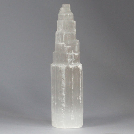 A tall, white selenite tower with stepped tiers stands upright on a gray surface. The crystal's texture shows vertical lines and subtle irregularities.