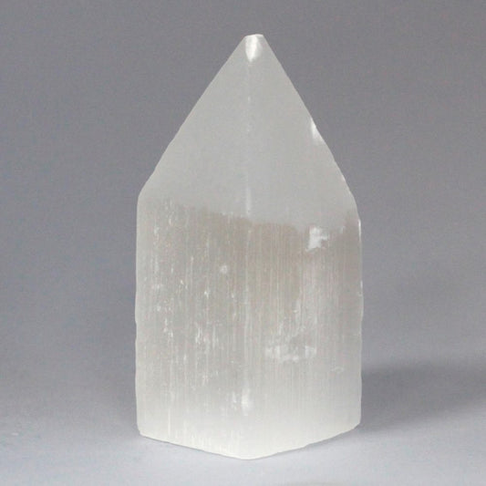A translucent selenite tower stands upright. Its lower portion is a slightly irregular rectangle, and its top tapers to a point. It is set against a gray background.